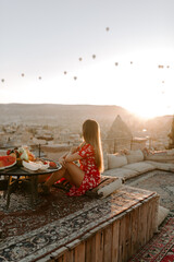 A young female traveller in a red dress is having early morning breakfast and watching hot air balloons in Cappadocia, Turkey on a hot summer day