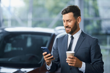 Handsome caucasian businessman drinking coffee and using modern smartphone while buying new car at salon. Concept of people, technology and transport.