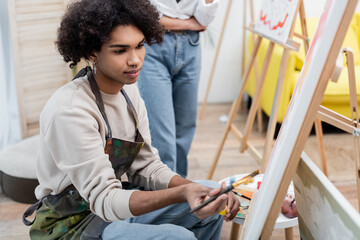 Young african american man painting on canvas near blurred girlfriend at home.