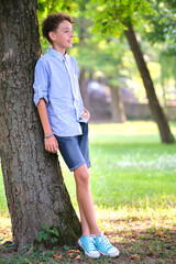 Young happy child boy relaxing in summer park leaning on tree trunk. Child wellbeing concept