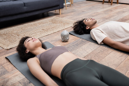 Young african american woman lying on yoga mat near incense stick and boyfriend meditating at home.