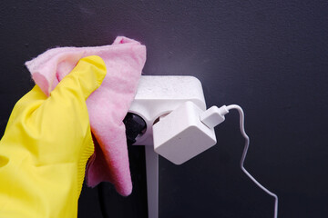 A woman's hand in yellow gloves wipes the dust with a pink cloth from a white electrical...