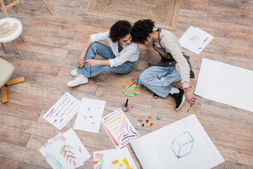 Overhead view of cheerful african american couple holding paintbrushes near papers and paints on floor.