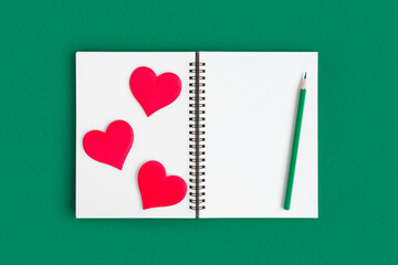 Notebook with pencil, red hearts. Open notebook on green emerald color background, spiral notepad. Top view. Notepad with blank white page for write, office flat lay. Copy Space. Valentine Day, Love