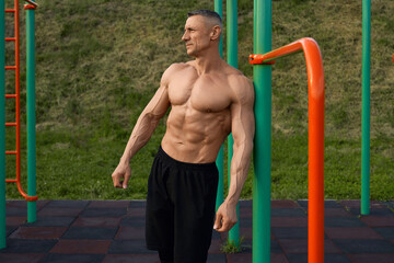 Strong caucasian bodybuilder in only shorts leaning on sport bars while standing outdoors and looking aside. Regular workout on sports ground. Active lifestyles.