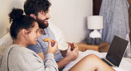 Date night done right. Shot of a happy young couple using a laptop and eating pizza while relaxing on the bed at home.