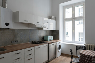 Interior of kitchen with large window in loft apartment. White modern furniture with cabinets and wooden counter. Dining table and wooden floor.