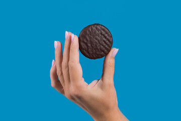 Close-up view stock photography of woman holding in between fingers small round brown object. Female hand with chocolate cookie isolated on blue background
