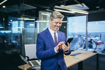Senior boss business man in modern office, portrait of business owner using phone smiling and rejoicing