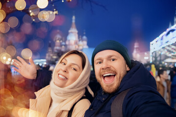 Happy friends man and woman making selfie photo and walking in night city Christmas market, bokeh...