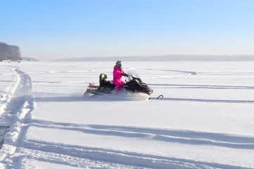 Fotobehang A girl rides a snowmobile through a snow-covered frozen lake. Riding on winter modes of transport. Active recreation. Snowmobiling in winter. Selective focus © Иван Решетников