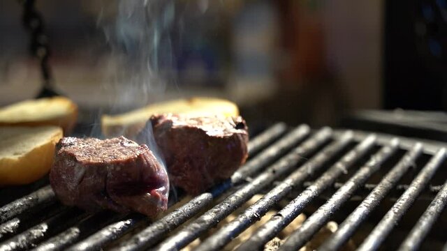 Filet mignon is fried on the grill close-up. In the background, cooking tongs are flipping a white bun.