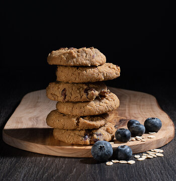 Oats cookies with blueberries on wooden chopping board and black background.