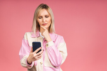 Fototapeta premium Blonde young female with terrible expression in smartphone, reads shocking news on web page, isolated on pink background. Shocked amazed woman using mobile phone.