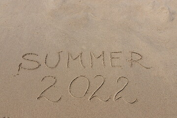 The inscription summer 2022 on the sand by the water and the rising wave, seashore beach vacation by the sea
