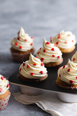 Vanilla muffins with buttercream frosting sprinkled with red sugar hearts in a muffin pan with a white piece of cloth, on a grey background. 