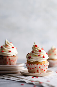 Vanilla muffins with buttercream frosting sprinkled with red sugar hearts on plates and a white cloth, on a grey background. 