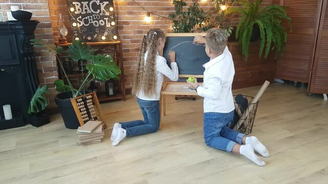 Blond schoolchildren - a boy and a girl draw a rainbow with multi-colored crayons on a blackboard against the background of fresh flowers, a brick wall and the inscription - Back to school!