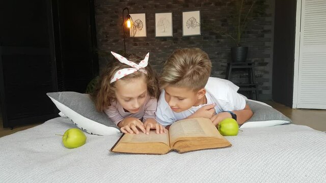 Children - a boy and a girl lie on the bed with a large encyclopedia and apples, read, talk. Home schooling.