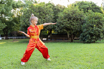 Fototapeta premium Cute little caucasian girl seven years old in red sport wushu uniform exercising in park at summer day. Lifestyle portrait of kung fu fighter child athlete