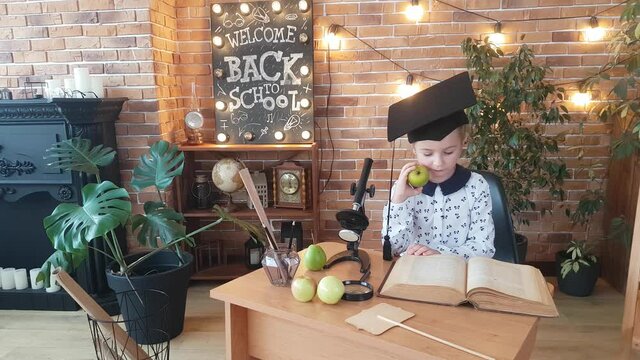 A schoolgirl in an academic square cap sits at a desk with a microscope and studies an encyclopedia while eating a pear. Background - a brick wall and the inscription Welcome! Back to school!