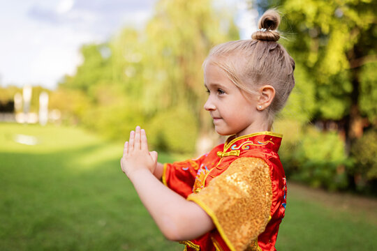Cute little caucasian girl seven years old in red sport wushu uniform exercising in park at summer day. Lifestyle portrait of kung fu fighter child athlete