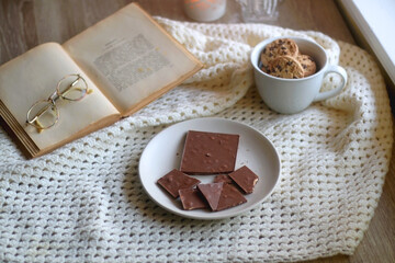 Plate with almond chocolate bar, mug with chocolate chip cookies, open book, reading glasses, soft blanket and lit candles. Hygge at home. Selective focus.