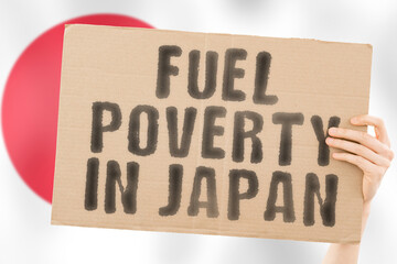 The phrase " Fuel poverty in Japan " on a banner in men's hand with blurred Japanese flag on the background. Economic. Expensive. Depressed. Gasoline. Fossil. Problem. Supply. Social issues