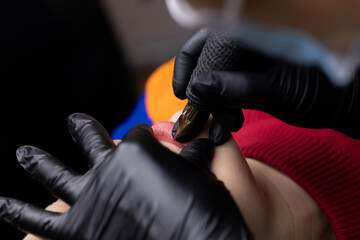 the permanent makeup artist in black sterile gloves bent over the model and performs the lip tattoo procedure - obrazy, fototapety, plakaty