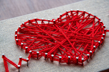Craft string art in shape of heart. Red woolen heart, symbol of love, made of red wool yarn threads tangled over metal nails on canvas background