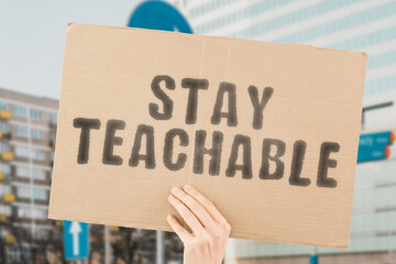 The phrase " Stay teachable " on a banner in men's hand with blurred background. Communicate. Educate. Personal improving. Creative thinking. Progressive. Progressing