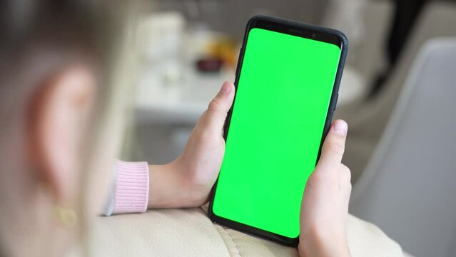 Little girl use smart phone with blank green screen mock up display for advertising text while rest on couch in living room at modern home. Phone with green screen and chroma key for copy space