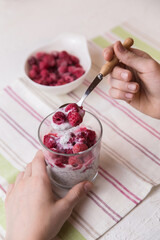 The hands of the child hold a glass with a dessert of yogurt, chia seeds, frozen raspberries