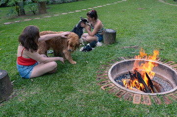 Two women sitting on the grass in the park around a firepit, on a bright afternoon, petting their...