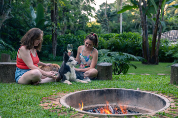 Two women sitting on the grass, drinking rosé wine in the park around a firepit, petting their Golden Retriever and another black and white mixed breed dog, during a bright warm afternoon. 