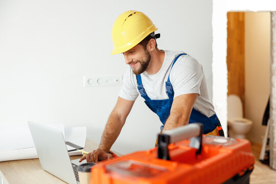 Smiling repairman in uniform and hard hat using laptop, looking through details of an order, preparing for renovation work
