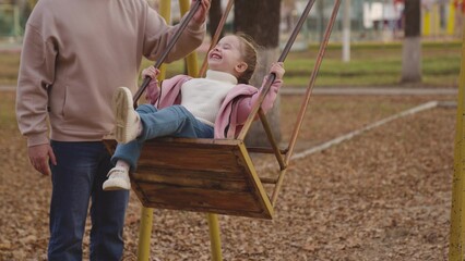 Fototapeta na wymiar mother shakes little child on playground swing, cheerful kid flies up and down, baby laughs and smiles while playing, mother and daughter on walk in city park, happy family, childhood dream of flying