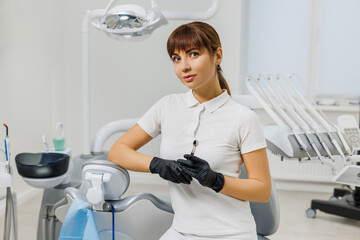 Woman dentist in white uniform and gloves sitting in stomatology cabinet among modern dental equipment. Portrait of female doctor - professional stomatologist, orthodontist.