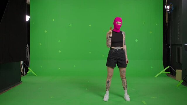 Young woman in pink balaclava with baseball bat in hands. Hooligan girl in mask walking and looking at camera on green screen background . Chroma key . 4k uhd video