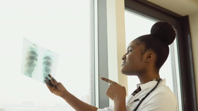 Female african american doctor or intern examines x-ray of lungs, holding it in hands indoors. Specialist holds transparent image of chest in arms, and carefully researches it
