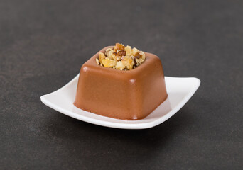 Small Chocolate cream dessert in the shape of a square savarin with a filling of crushed nuts on a serving plate on a dark gray background