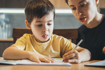 Boy doing his homework with his mother sitting at desk, at home, he is writing on copybook. Child...