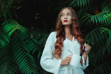 Obraz na płótnie Canvas Gorgeous girl in a white coat against the background of large green leaves. Cup and black glasses in hands. Red lipstick and arrows. Morning portrait 