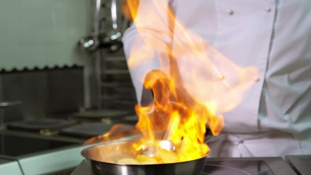A close-up of a professional chef in black gloves in the kitchen of a restaurant cooks in the style of Flambe. Fruits and citrus fruits in a frying pan with an open fire.Exquisite French haute cuisine
