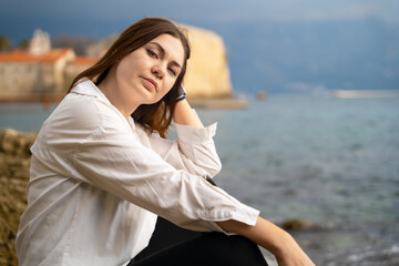 Active woman sits on beach in relaxed pose and adjusts her hair with hand, blurred background. Modern business lady is resting on sea resort during vacation
