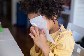 Little mixed race girl cleaning her nose with a tissue after sneezing because of COVID19 variant...