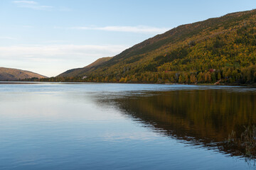 A wide river with smooth blue water. There is a luscious green, red and golden forest of trees along the riverbank.  The calm and peaceful pond water is reflecting the sky and autumn landscape.