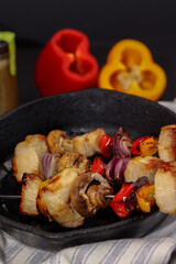 chicken meat skewers with purple onion, red, orange and yellow peppers on iron pan with black background and white striped napkins