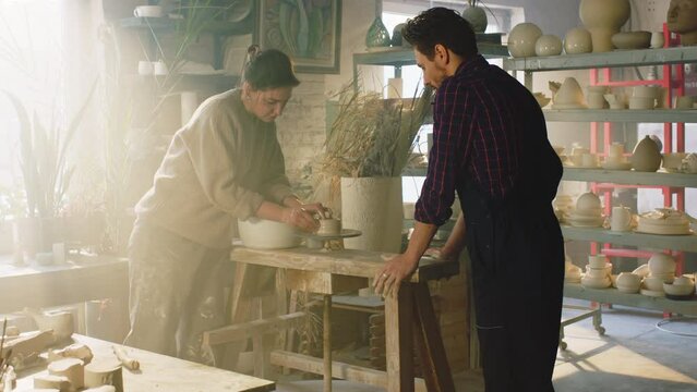 Young woman, potter, is giving masterclass to man, showing how to work with raw clay on pottery wheel, demonstrate how to model small pot, Slow motion.