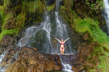 Full view of caucasian woman in swimsuit at thermal springs in Colombia. Horizontal panoramic view of woman raising up arms underneath a big waterfall in the jungle. Colombia travel destination